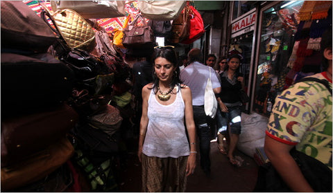 A woman wearing, Kiran Rai, a white tank top and patterned skirt walks through a crowded, narrow market street filled with shoppers and hanging bags. 17 Nov 2009, “Even In a Terror Attack, Seeing the Best of India”